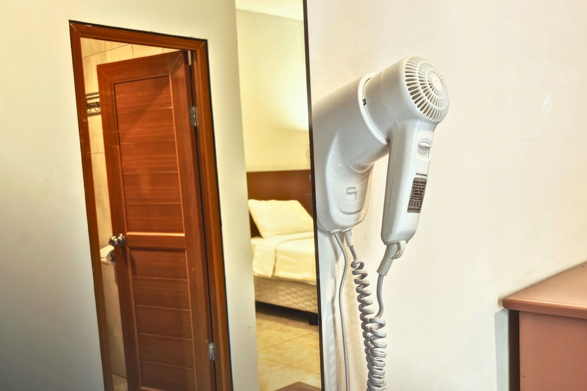 Une pièce avec une porte en bois, un lit avec du linge de lit blanc et un sèche-cheveux blanc fixé au mur.