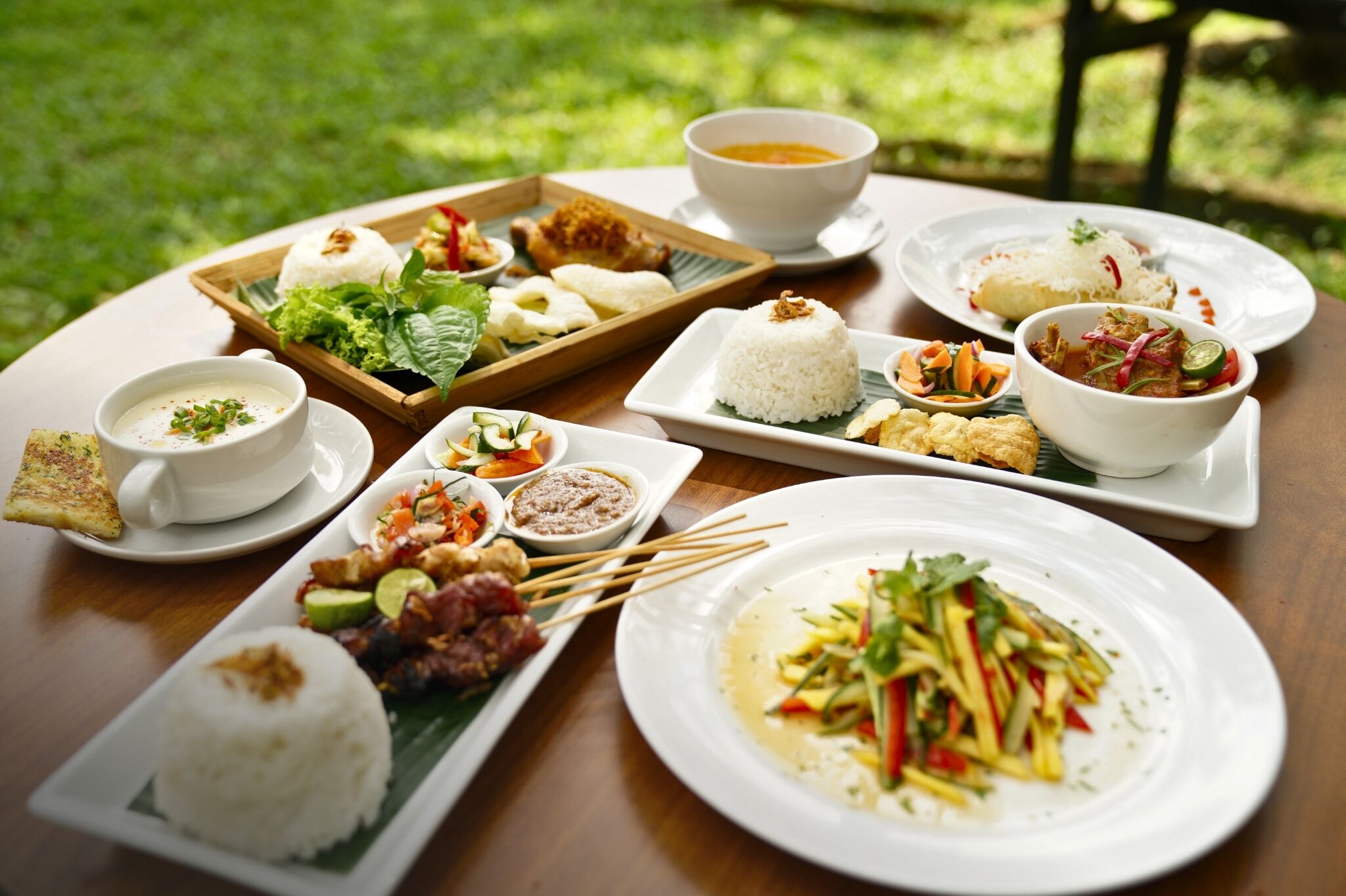 A table with various dishes including rice, soup, and a salad, set outdoors on a wooden table.