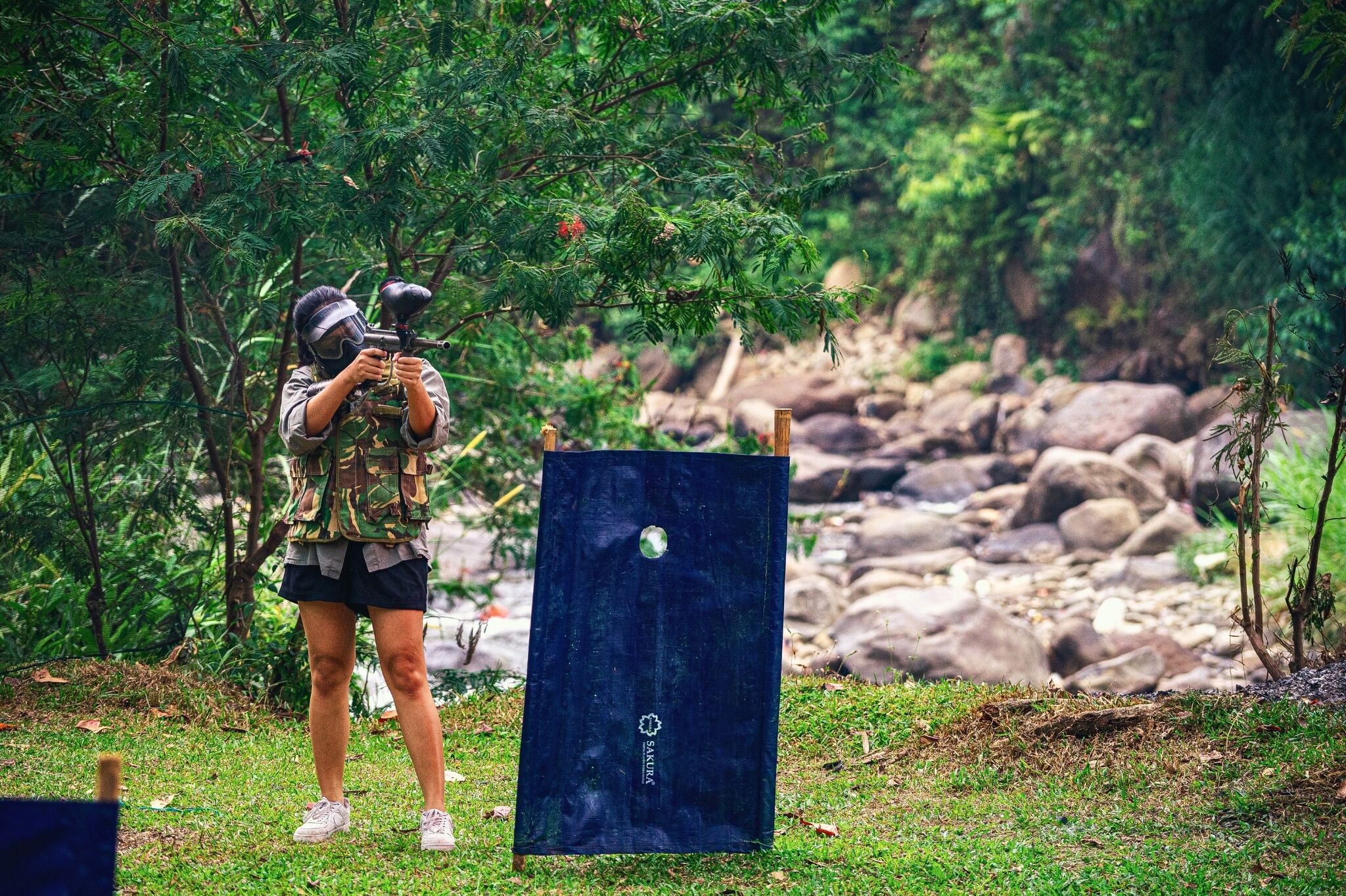 Una persona apuntando con un arma de paintball a un blanco en un entorno natural con rocas y follaje.