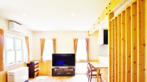 A living room with a flat-screen TV, a glass coffee table, a sofa, and a dining area with chairs.