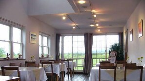 A dining area with tables set for guests, large windows offering a view of the outdoors, and a ceiling with recessed lighting.
