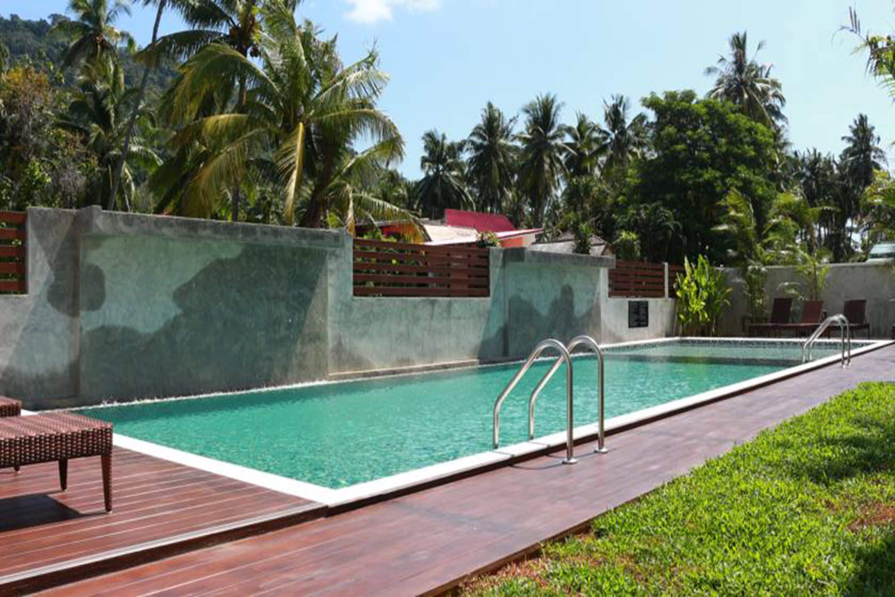 A rectangular swimming pool with a wooden deck and metal railings, surrounded by lush greenery and palm trees.