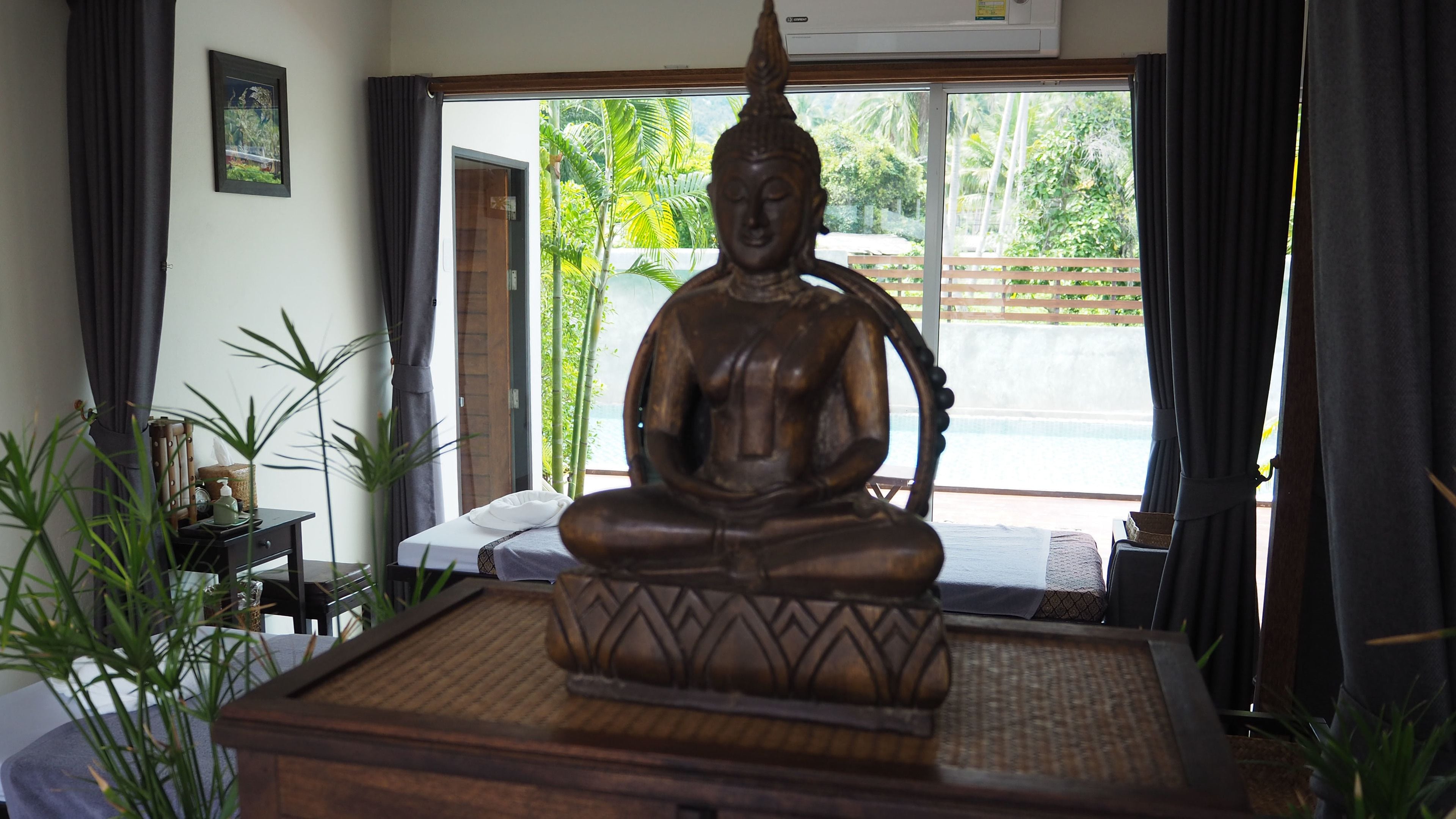 Una habitación con una gran estatua de Buda sobre una plataforma, una cama con colchón blanco, una mesita con una botella y una caja de pañuelos, y una ventana que ofrece vistas a zonas verdes y una valla.