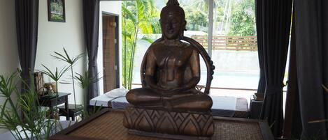 Una habitación con una gran estatua de Buda sobre una plataforma, una cama con colchón blanco, una mesita con una botella y una caja de pañuelos, y una ventana que ofrece vistas a zonas verdes y una valla.