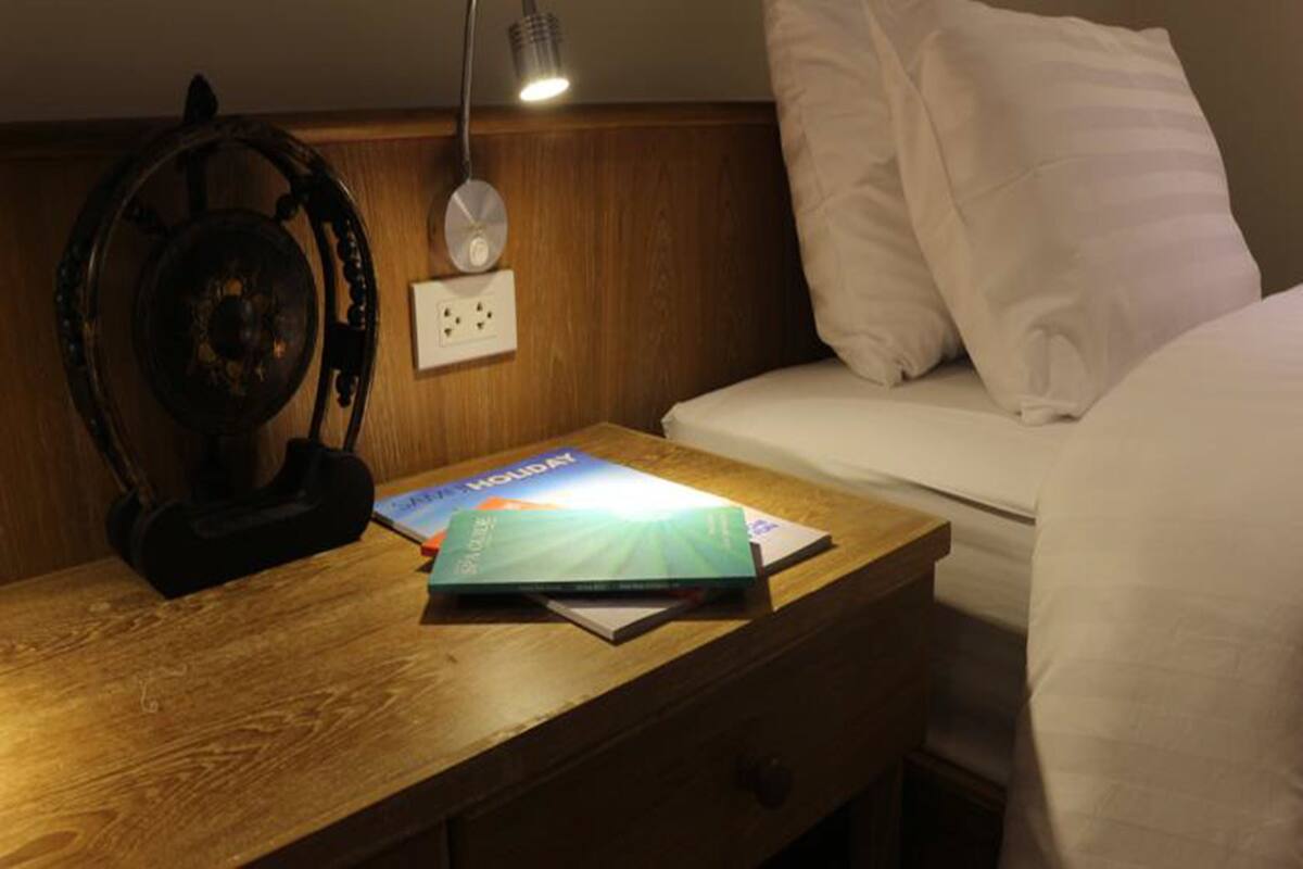 A neatly made bed with white linens, a wooden bedside table with a book and a lamp, and a wall-mounted power outlet.