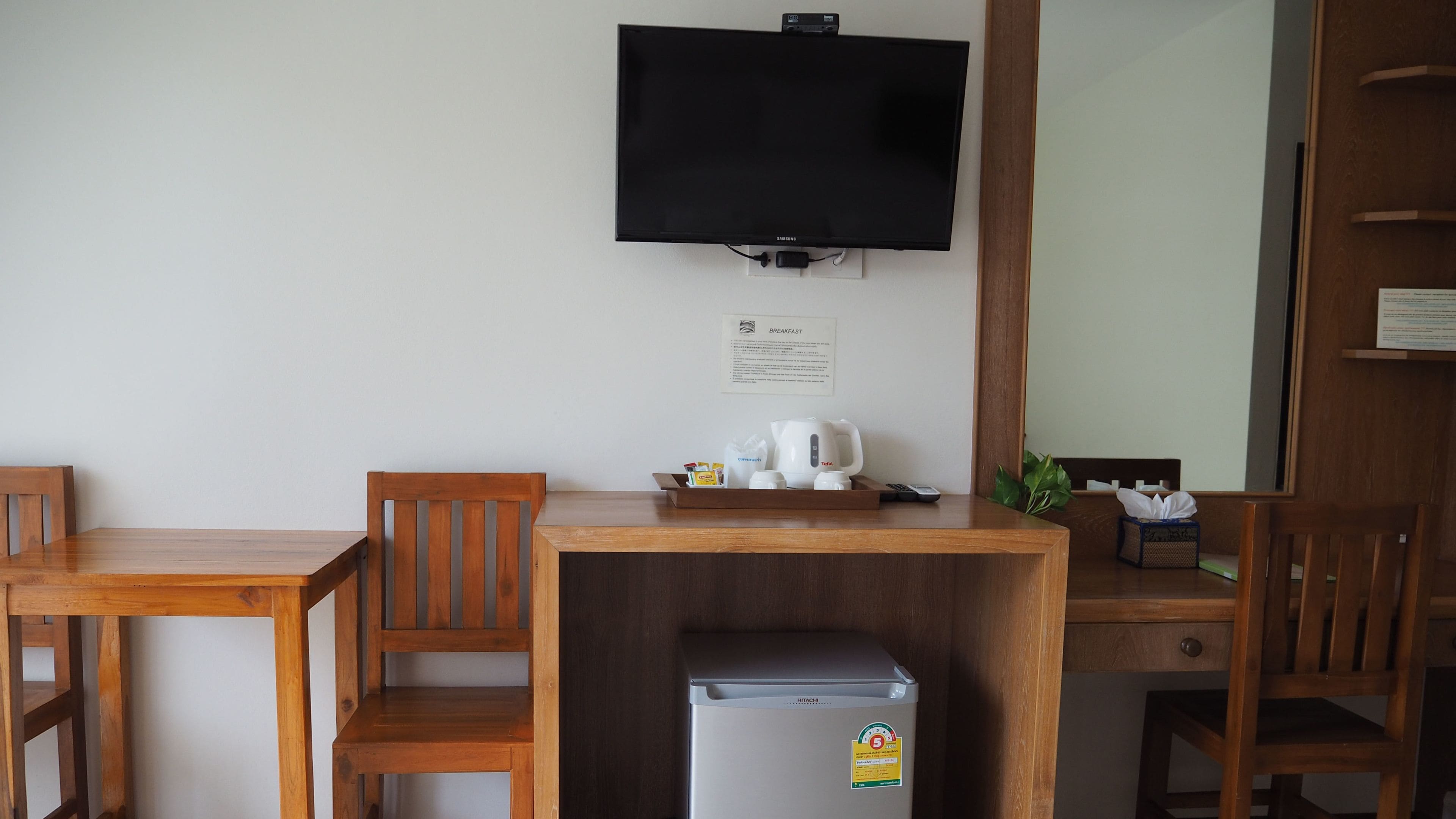 A hotel room with a wooden desk, a television mounted on the wall, a refrigerator, and a small plant.