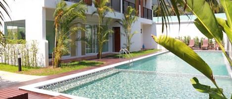 Una piscina con borde de azulejos, rodeada por una terraza de madera y un edificio blanco con balcones.