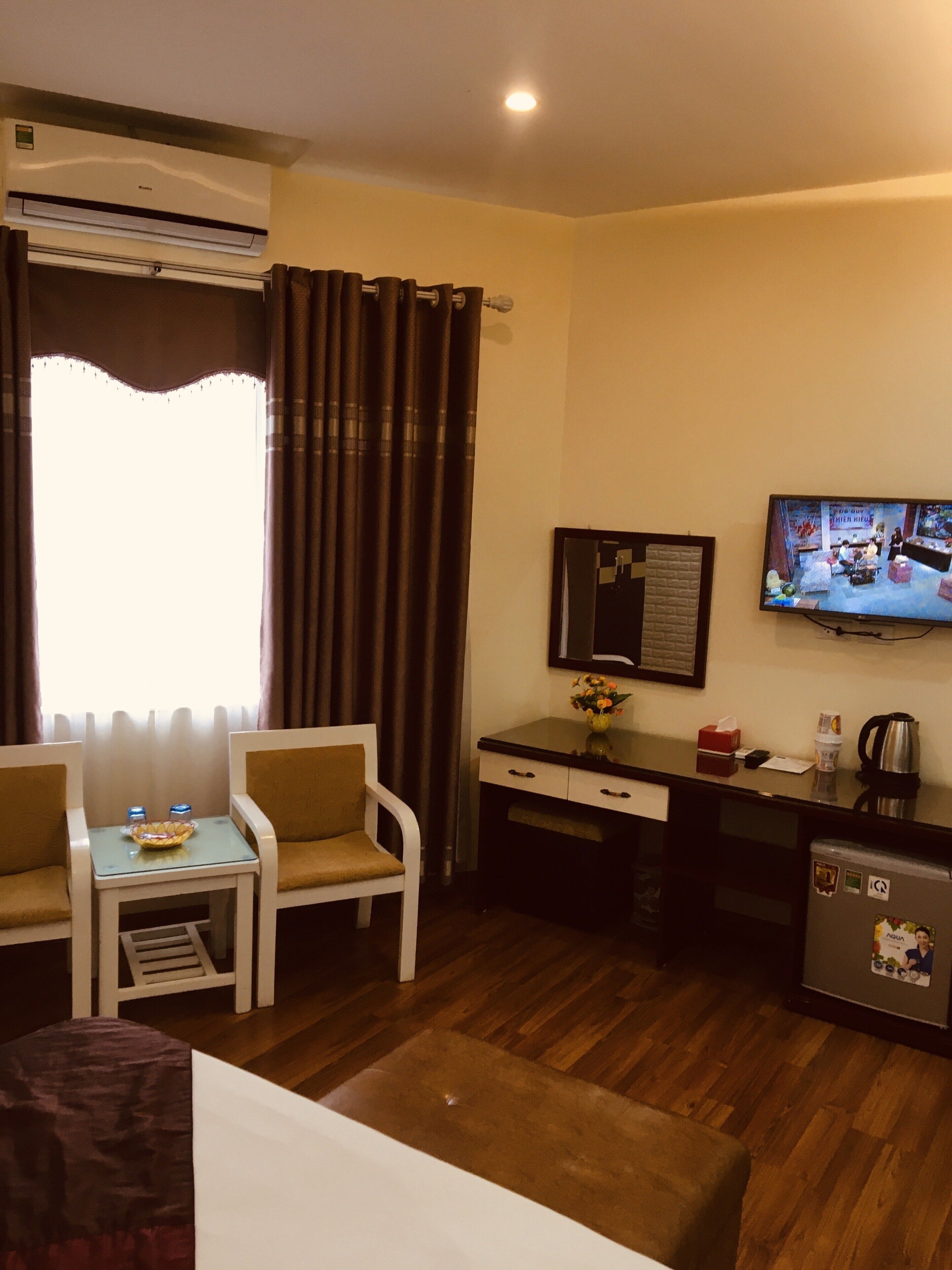 A hotel room with a TV, a desk, a chair, a small table, and a refrigerator.