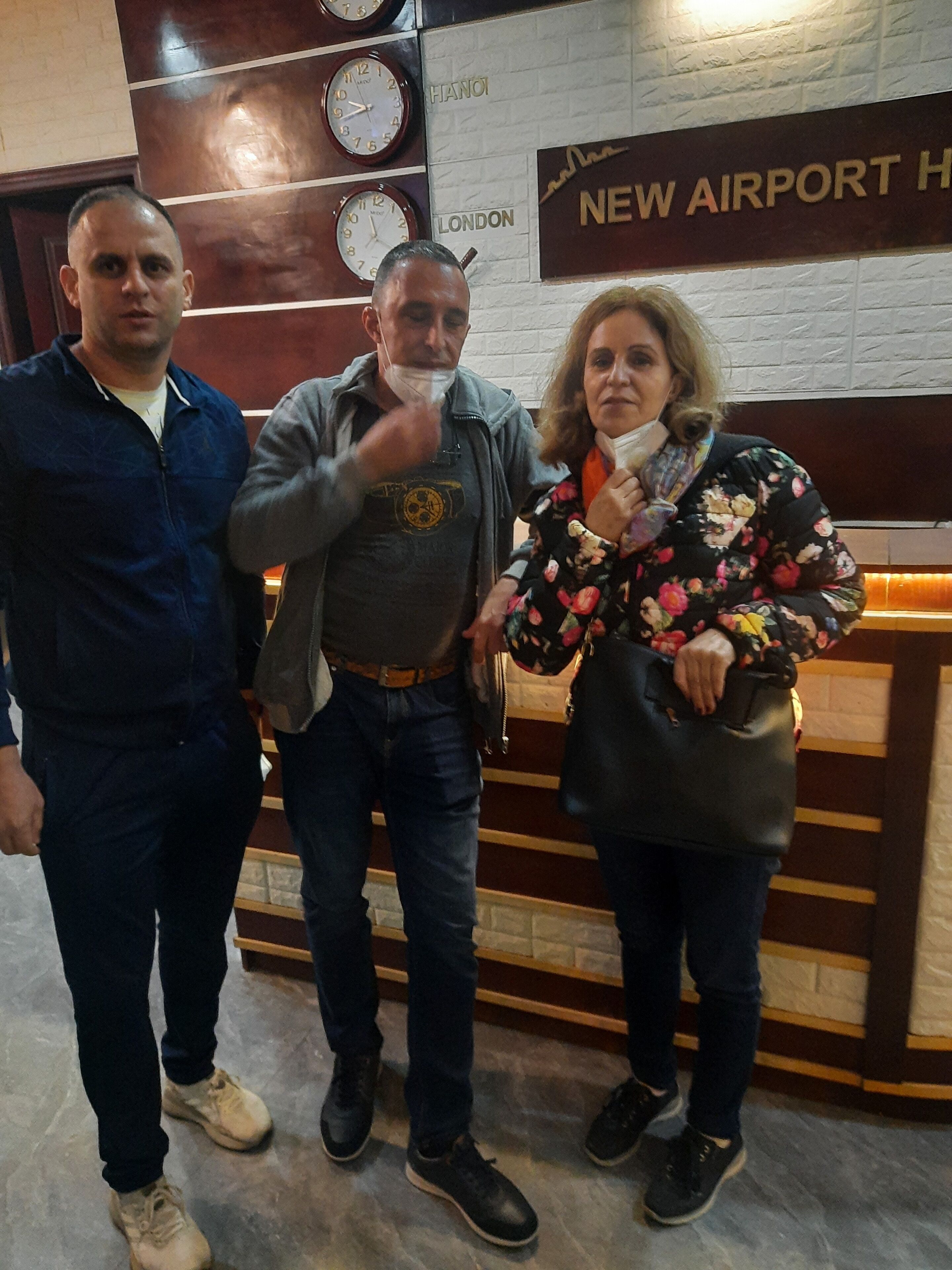 Tres personas frente a la mesa de recepción, con relojes y carteles que indican 'New Airport' y 'London'.