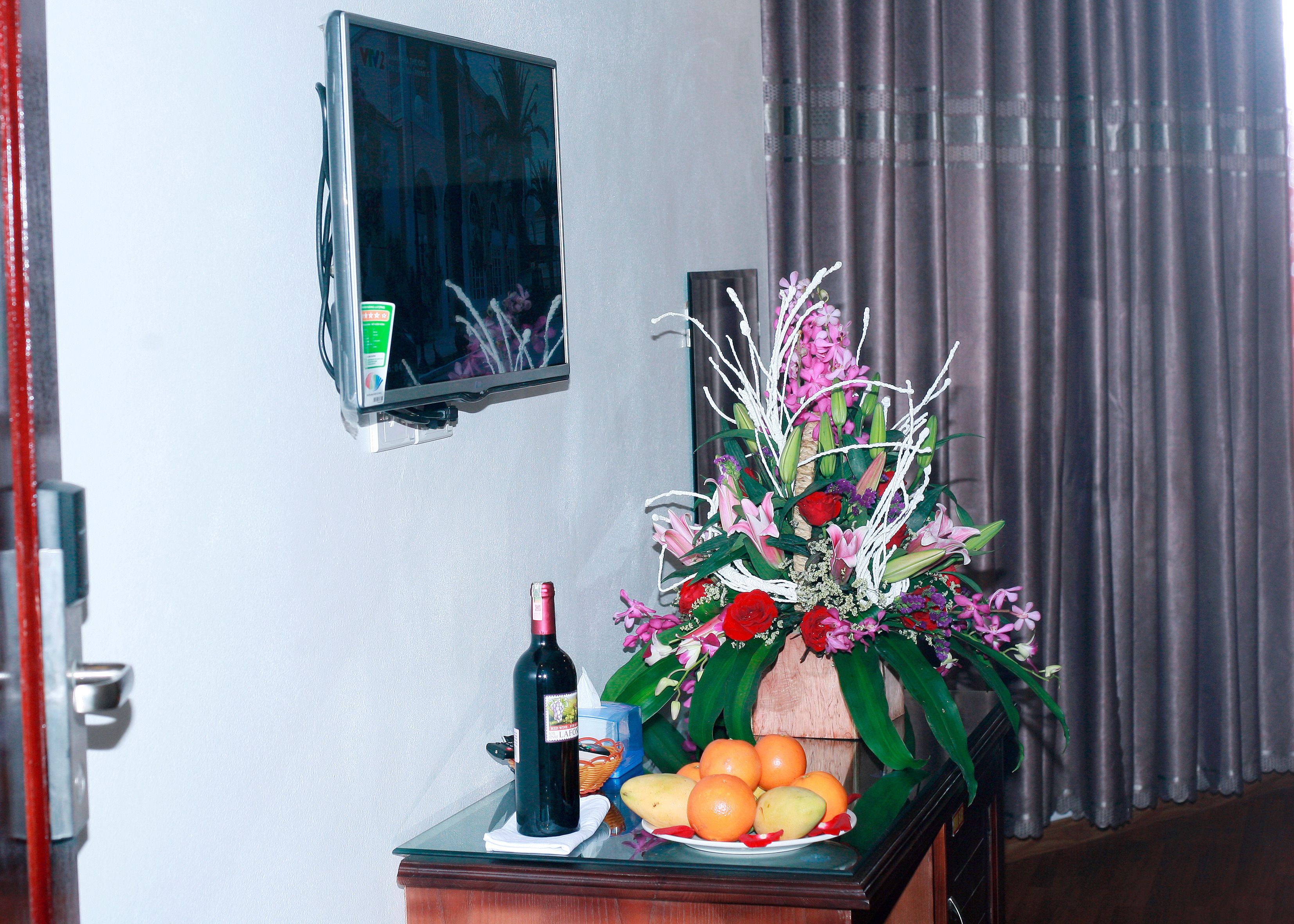 A room with a TV mounted on the wall, a table with a bottle of wine, a bowl of fruit, and a floral arrangement.