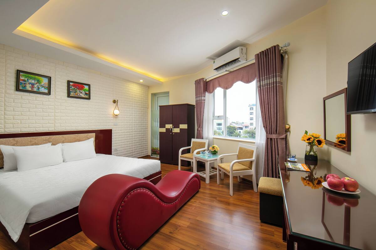 Quarto de hotel com cama, sofá vermelho, mesa de jantar com cadeiras, TV e janela com cortinas.