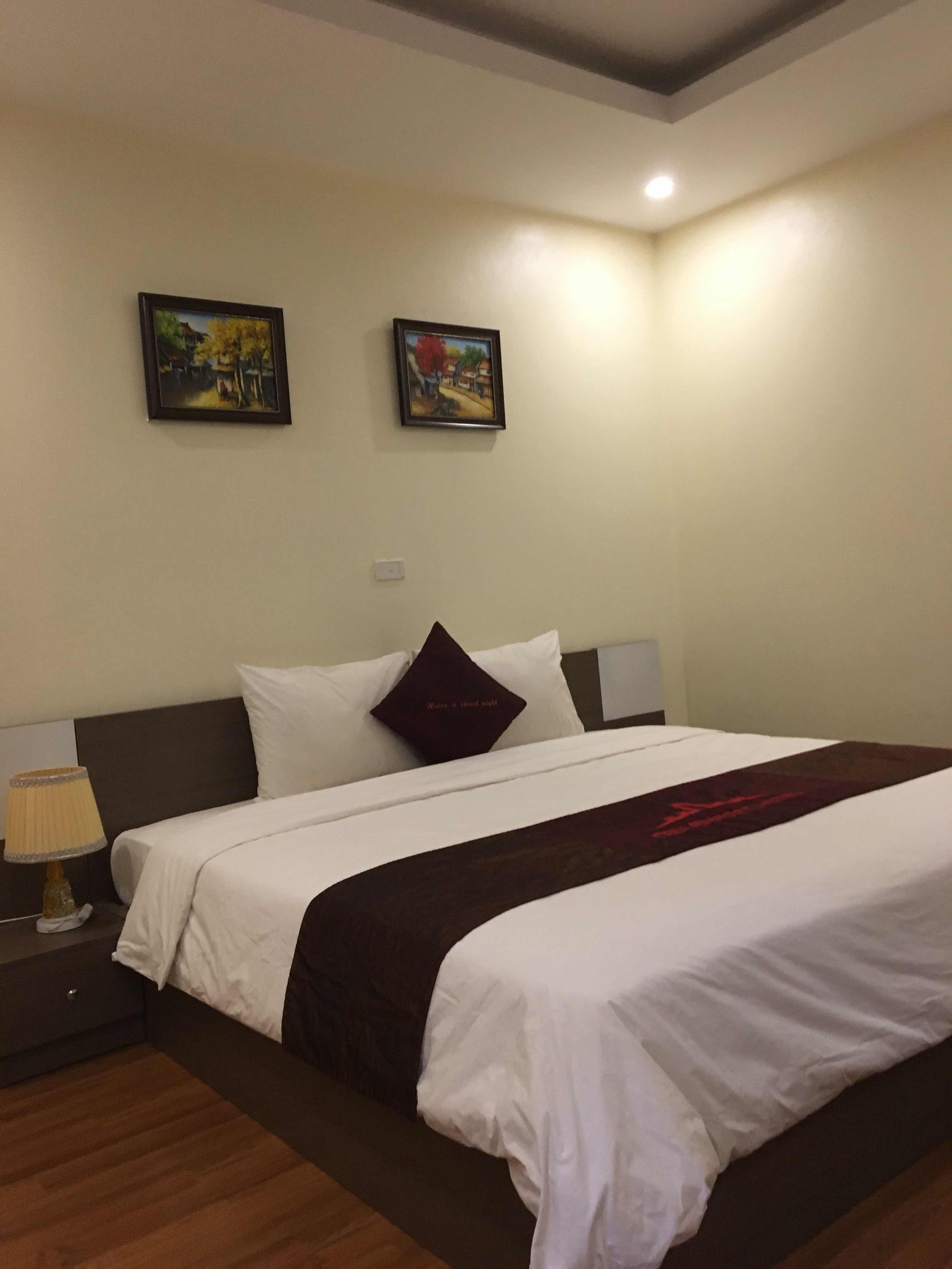Habitación de hotel con una cama grande, dos cuadros en la pared, una lámpara de noche y un cabecero de color oscuro.
