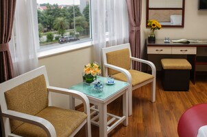 Quarto com duas cadeiras brancas com almofadas castanhas, uma mesa de vidro pequena, um vaso com flores e uma escrivaninha com um espelho e um vaso com girassóis.