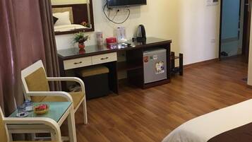 A hotel room with a bed, a desk with a computer, a chair, a small table, a TV, and a refrigerator.