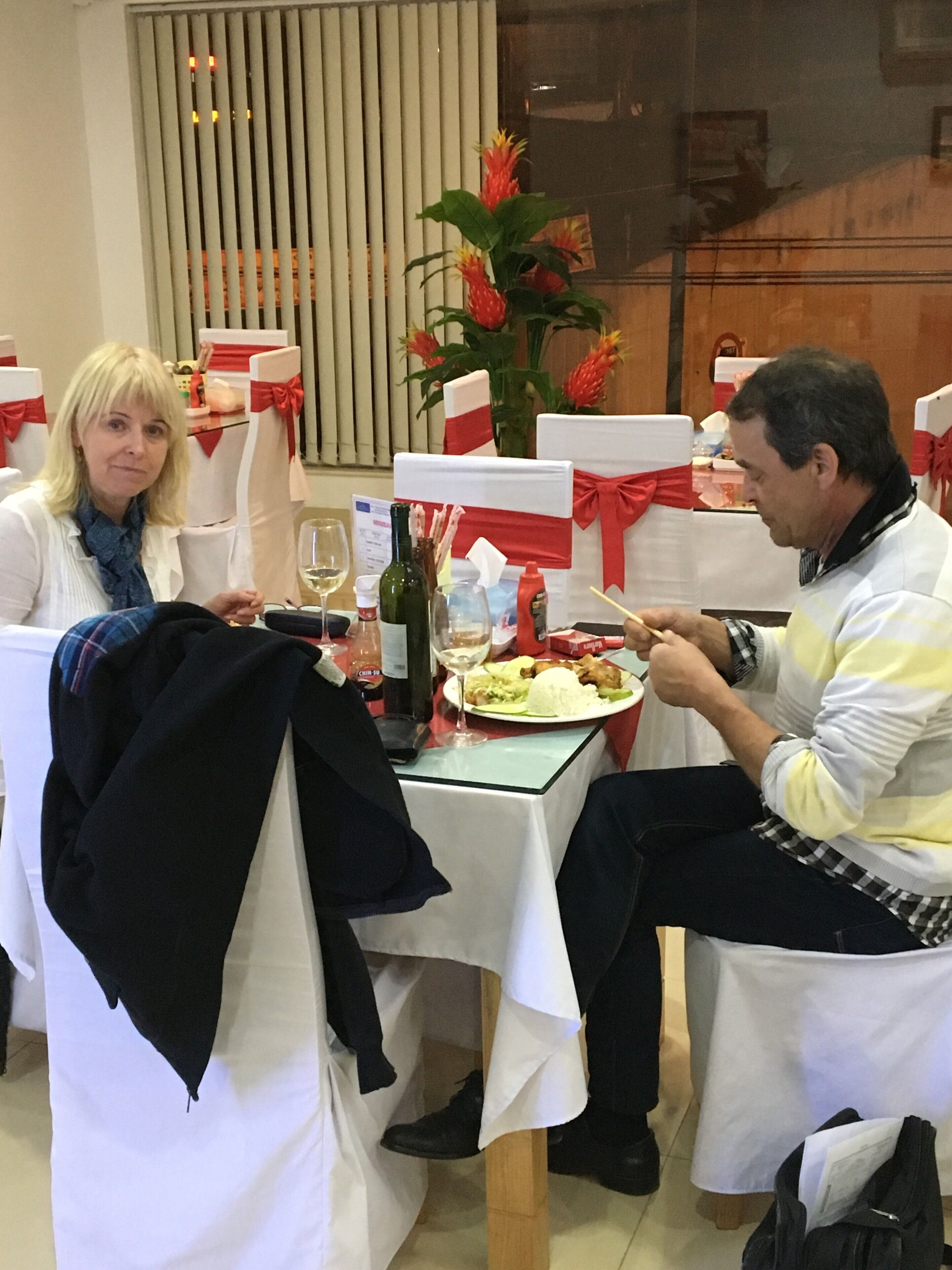 A dining table with a bottle of wine, a glass, and a plate of food. There are chairs with white covers and red ribbons. A plant with red flowers is on the table.