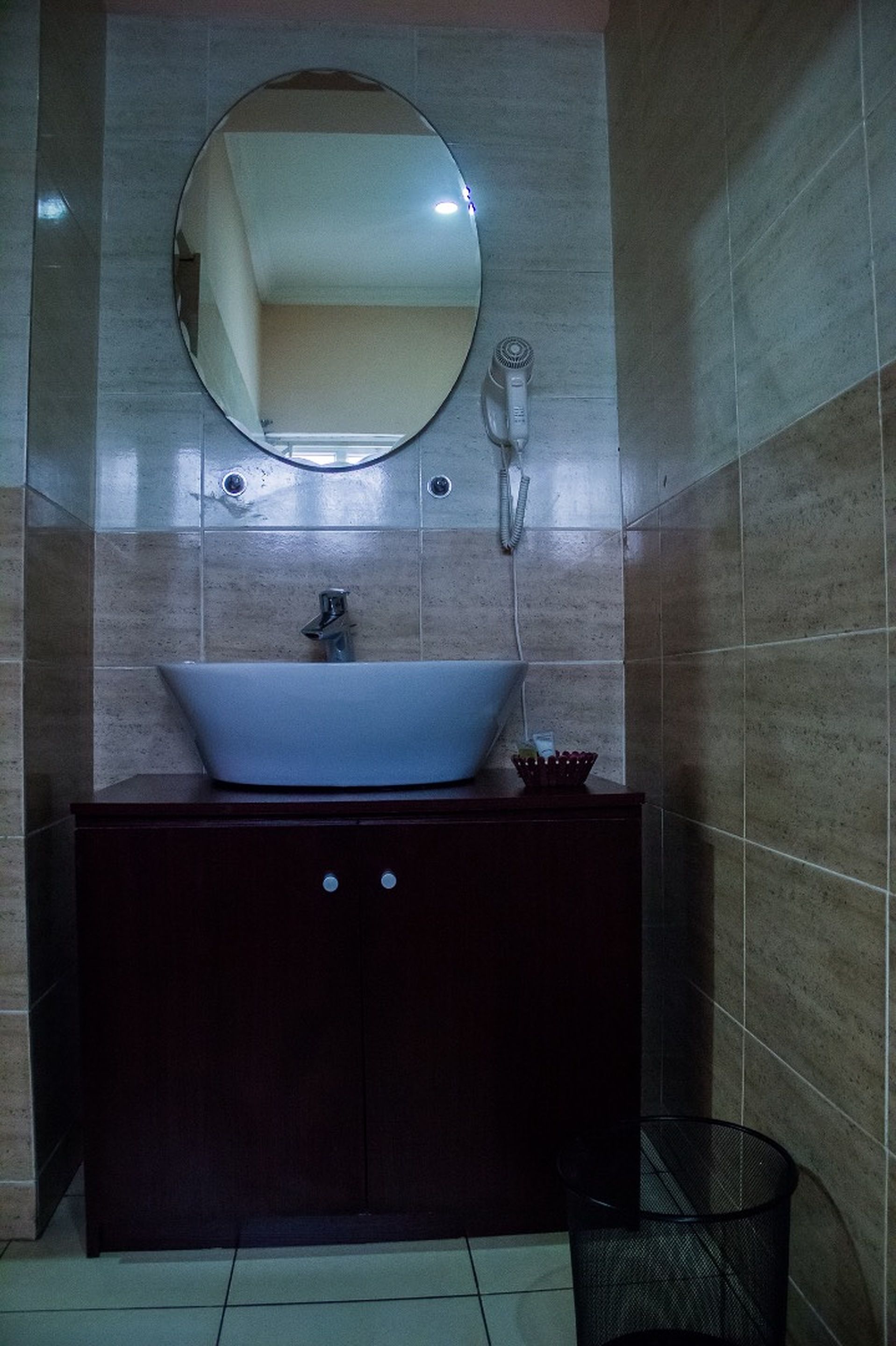 A bathroom with a white sink, dark wooden vanity, a round mirror, and a hairdryer on the wall.