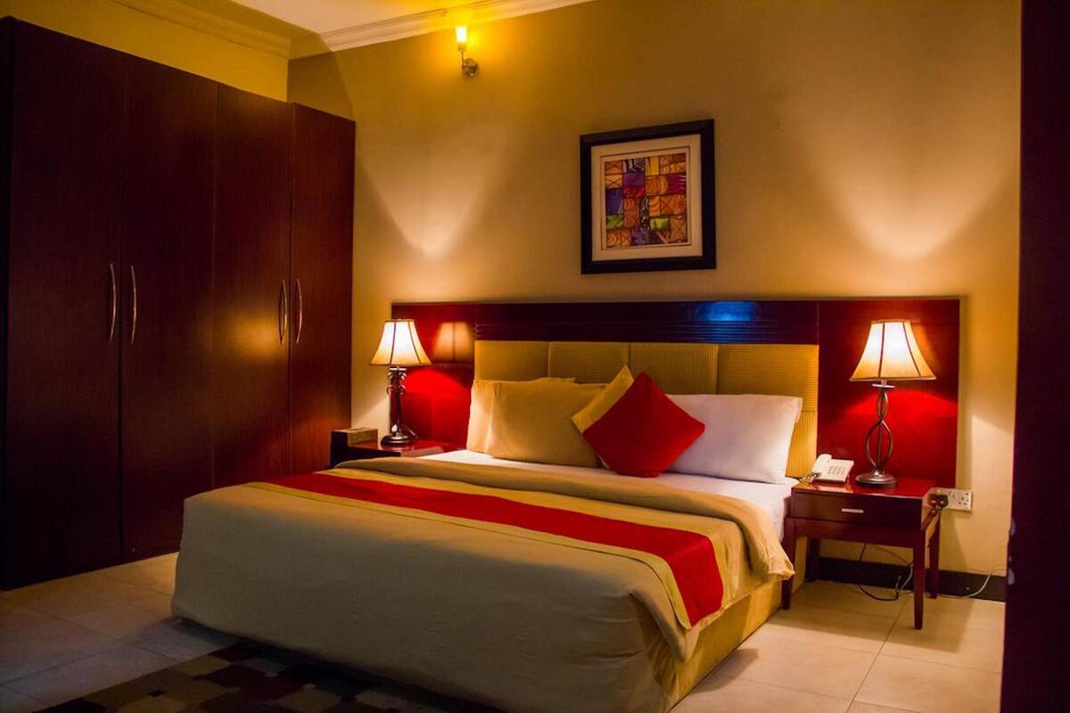 A hotel room with a large bed, two bedside tables, a nightstand, a wardrobe, and a framed artwork on the wall.