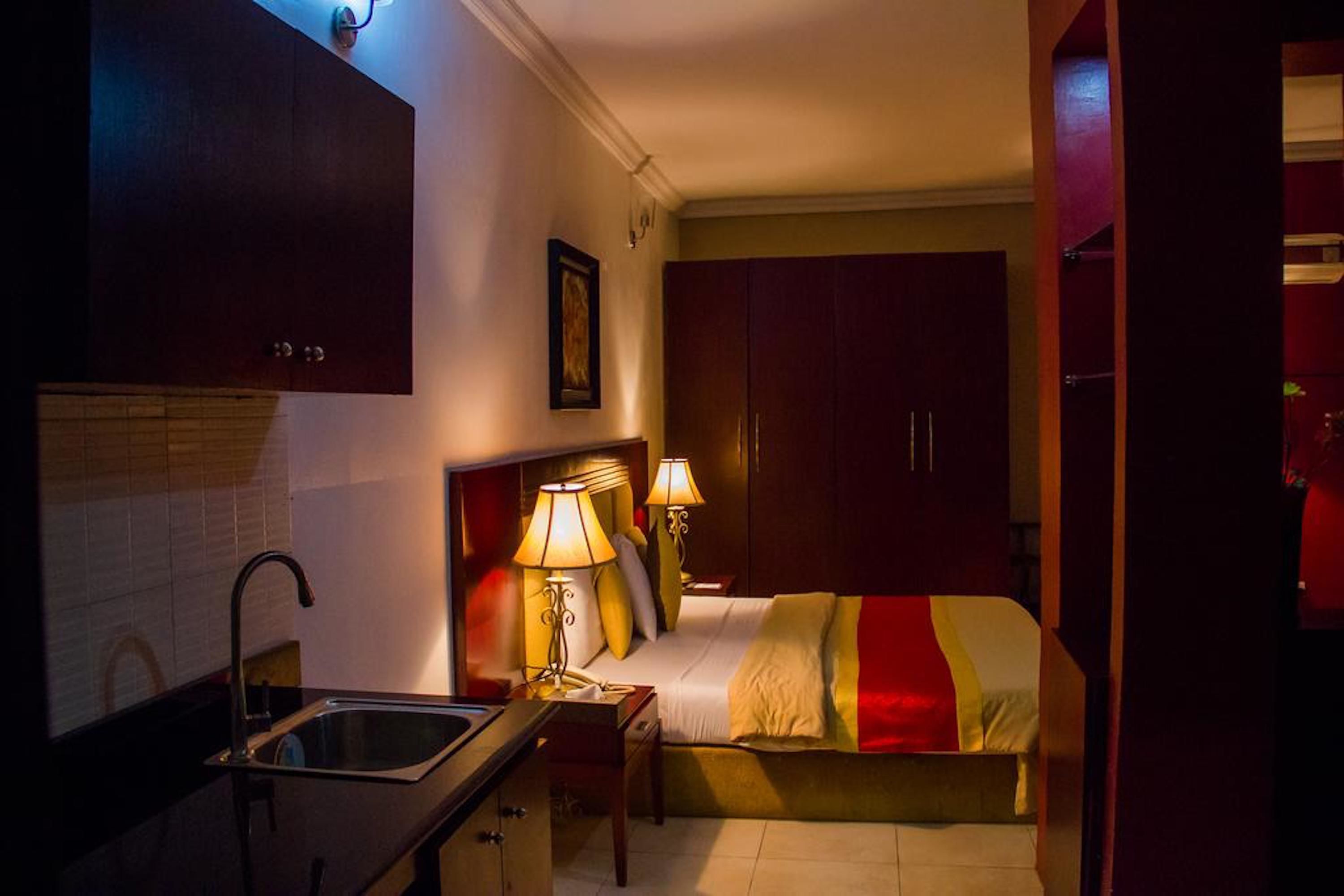 A compact hotel room with a kitchenette, a bed with a red and white striped blanket, and two bedside lamps.