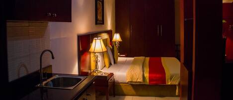 A compact hotel room with a kitchenette, a bed with a red and white striped blanket, and two bedside lamps.