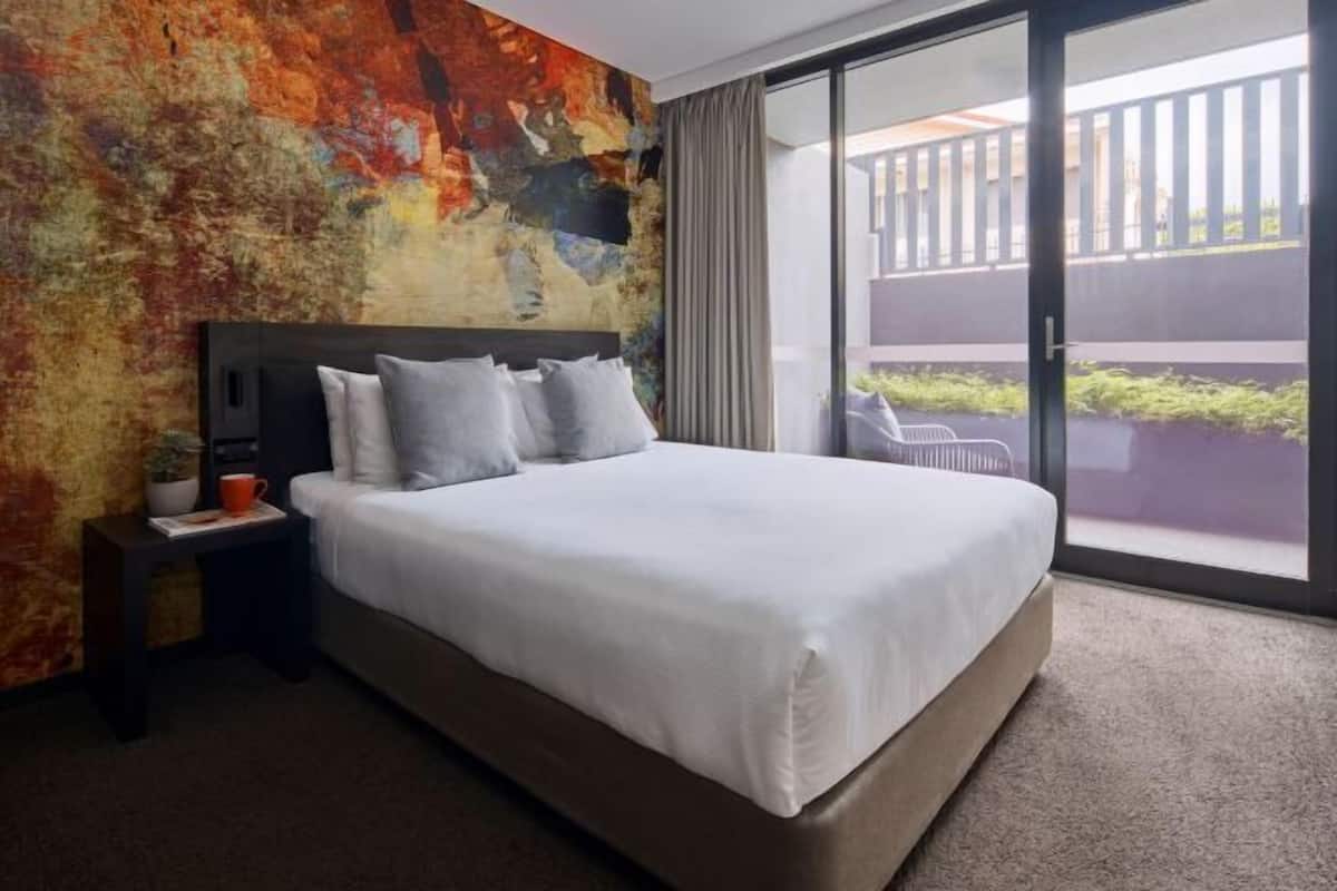 Ein modernes Hotelzimmer mit einem großen Bett, einem Nachttisch, einer farbenfrohen abstrakten Wand und einer Glastür zum Balkon.