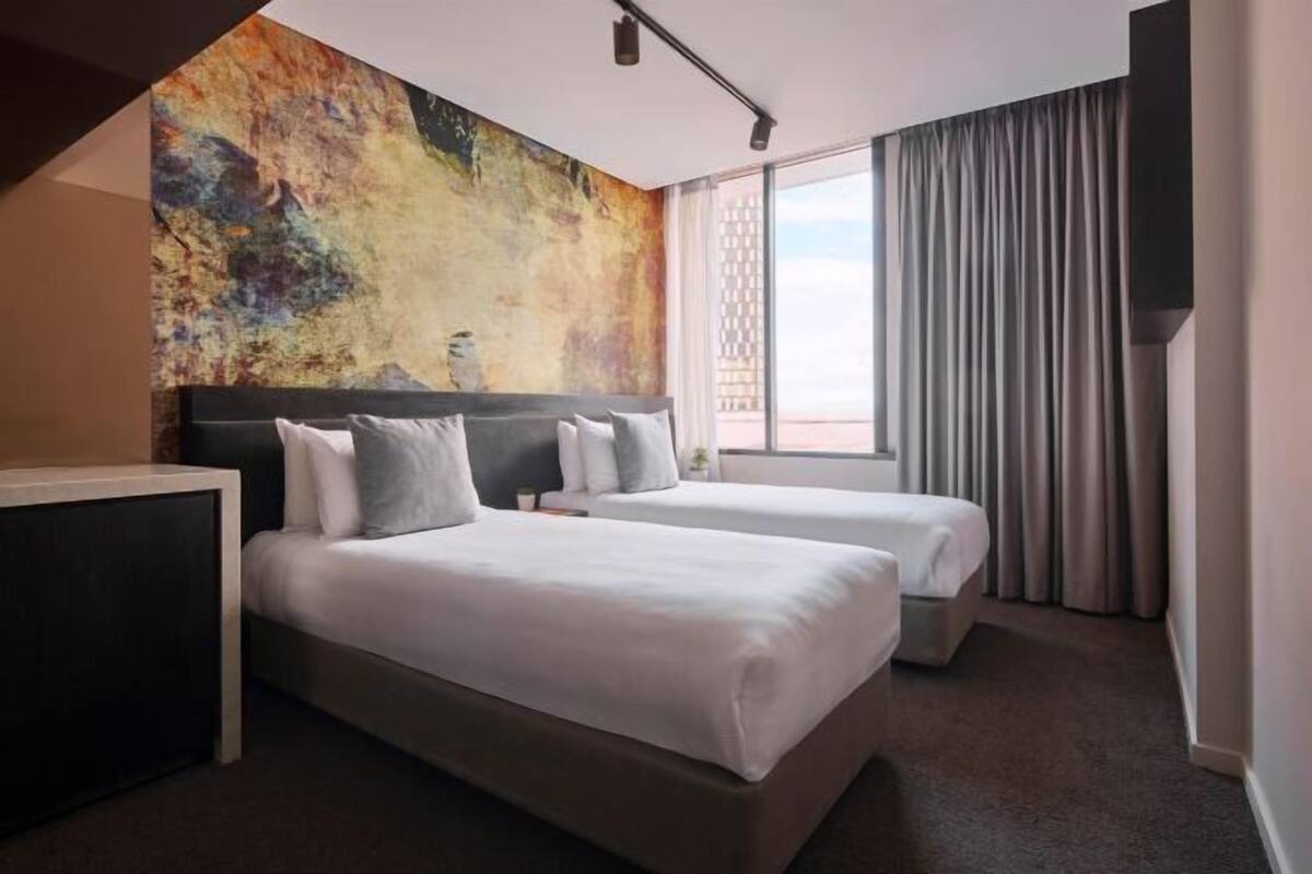 Ein Hotelzimmer mit zwei Betten, einem großen abstrakten Gemälde an der Wand, einem Fenster mit Vorhängen und Blick nach draußen.