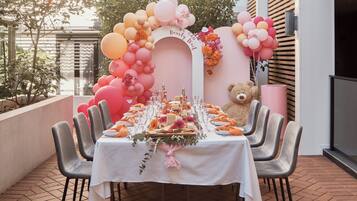 Un espace extérieur aménagé pour les repas, avec une table blanche dressée pour un repas, entourée de chaises. Un ours en peluche est posé sur la table, et un arrangement floral orne le mur.