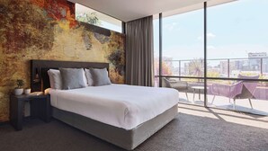 Una habitación de hotel moderna con una cama grande, vistas de la ciudad a través de ventanales del suelo al techo y un balcón con mobiliario exterior.