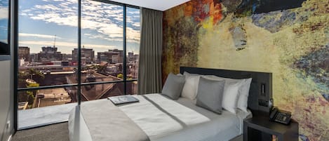 Ein modernes Hotelzimmer mit einem großen Bett, einem Nachttisch mit Telefon und Blick auf eine Stadtlandschaft durch raumhohe Fenster.