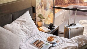 Una cama bien hecha con almohadas, un libro, una taza de café, un plato con un pastel y una maleta sobre una mesita de noche.