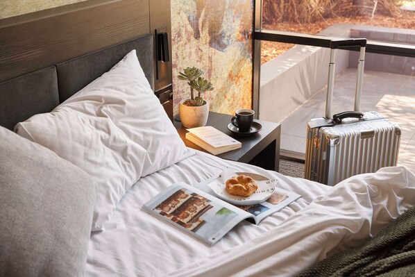 Una cama bien hecha con almohadas, un libro, una taza de café, un plato con un pastel y una maleta sobre una mesita de noche.