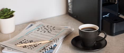 Una cafetera, una taza de café, un periódico y un bolígrafo sobre una mesa.