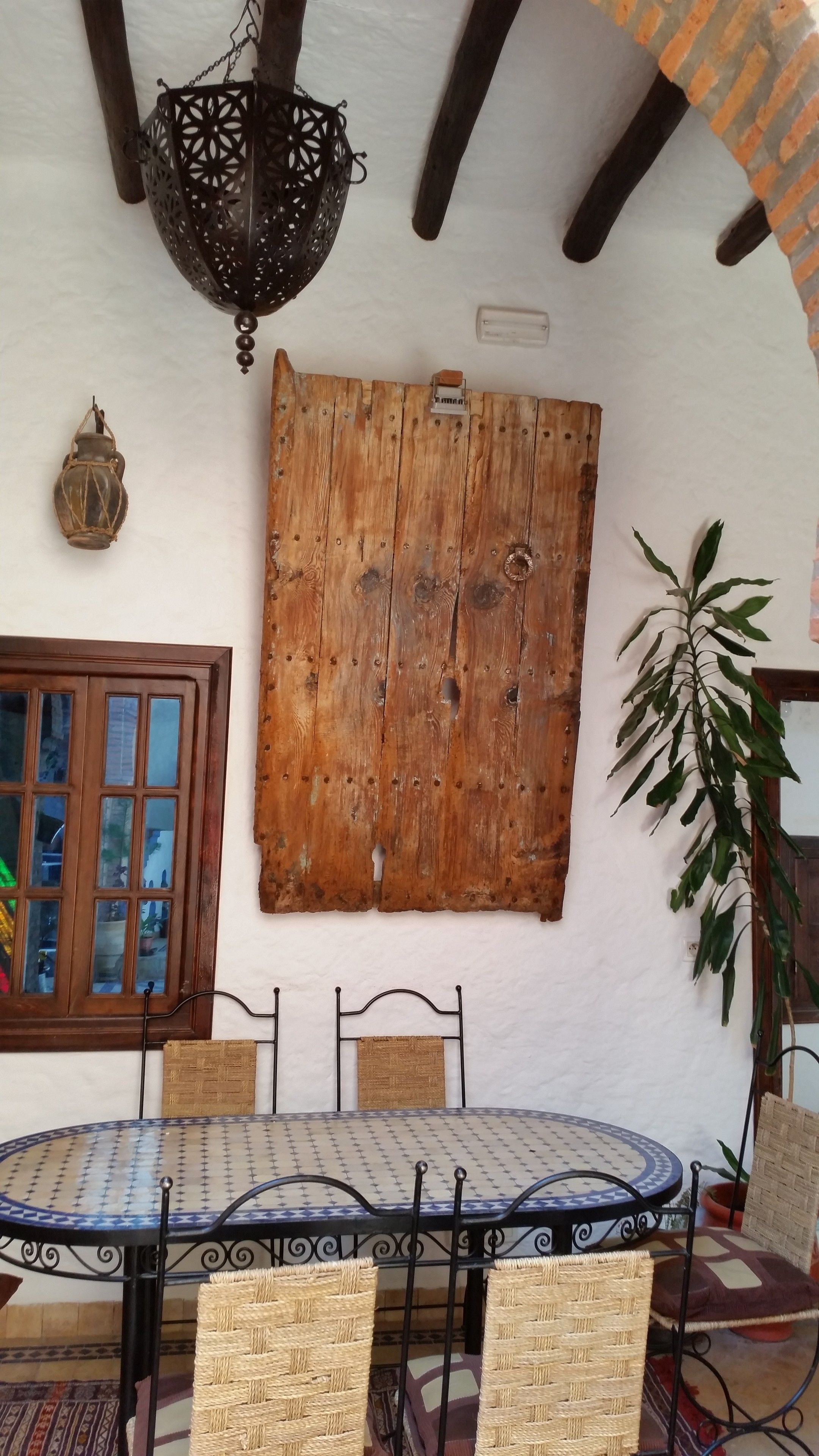 Un comedor rústico con una mesa redonda, sillas de mimbre, un panel de pared de madera, un farol colgante y una ventana con contraventanas de madera.