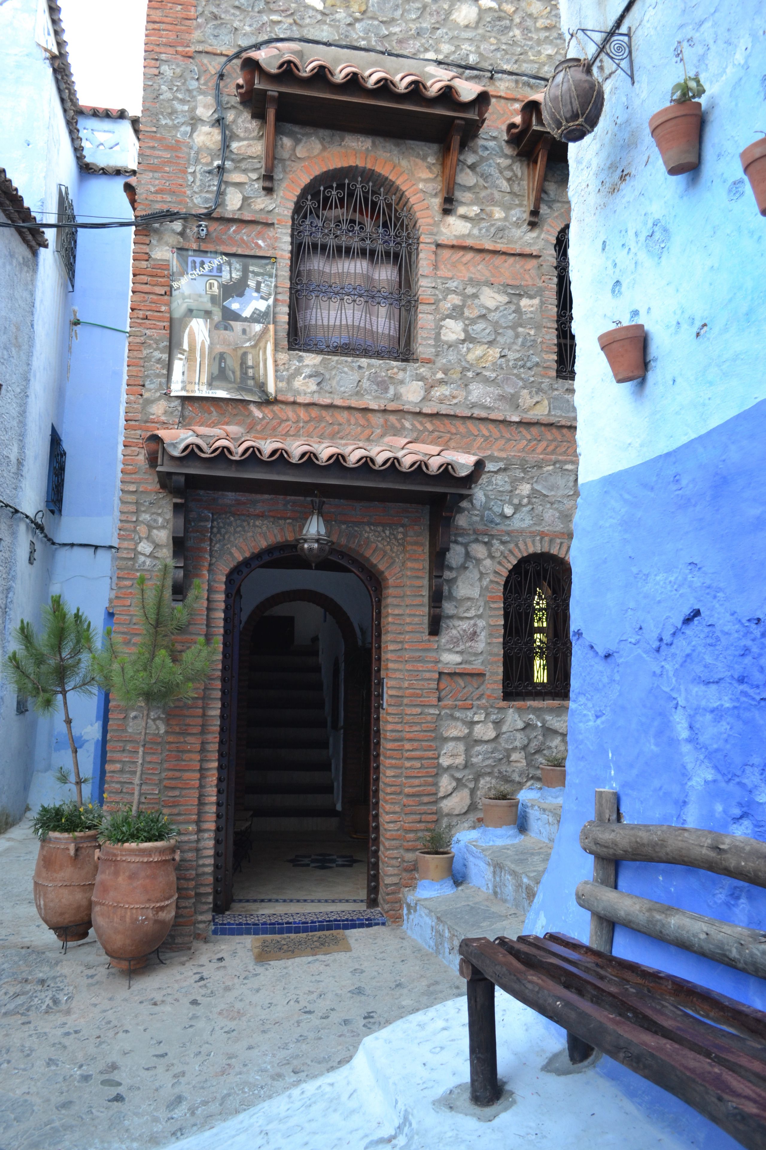 A narrow street with a stone building featuring an arched entrance, potted plants, and a wooden bench.