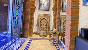 Un interior tradicional marroquí con piso de azulejos, una puerta de madera con panel de vidrio y una pared decorativa con un cuadro enmarcado.