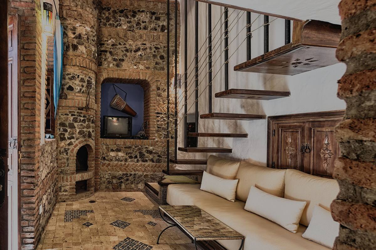 Uma sala de estar aconchegante com parede de pedra, escada de madeira e sofá bege.