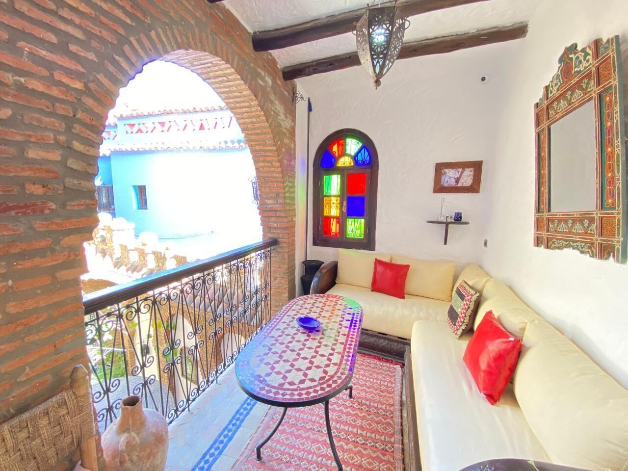 A balcony with a wrought iron railing, a small table with a bowl, a sofa with cushions, a colorful stained glass window, and a decorative mirror.