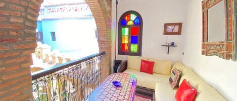 A balcony with a wrought iron railing, a small table with a bowl, a sofa with cushions, a colorful stained glass window, and a decorative mirror.