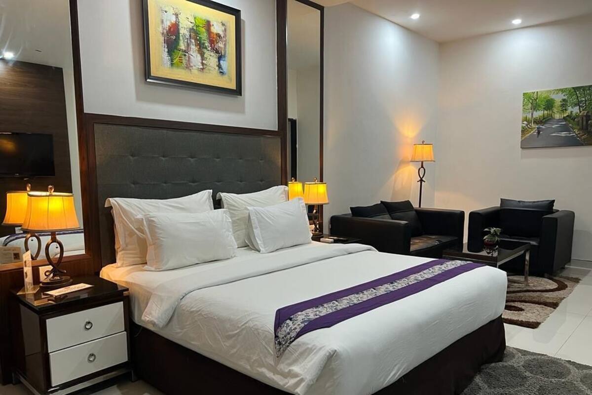 A modern hotel room with a large bed, a desk, two chairs, a TV, and artwork on the walls.