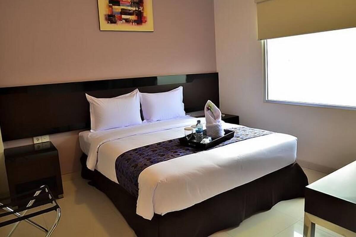 A hotel room with a bed, a nightstand, a chair, a window, and a painting on the wall.