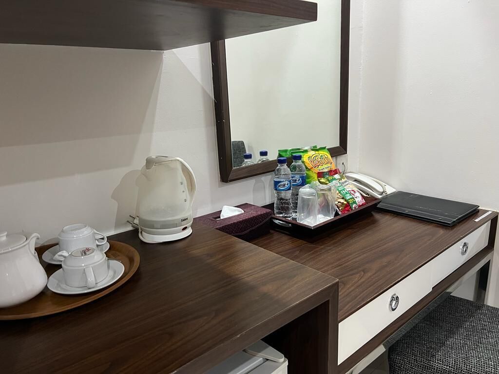 A hotel room with a wooden desk, a white kettle, a tray with cups, a mirror, and various snacks and drinks.