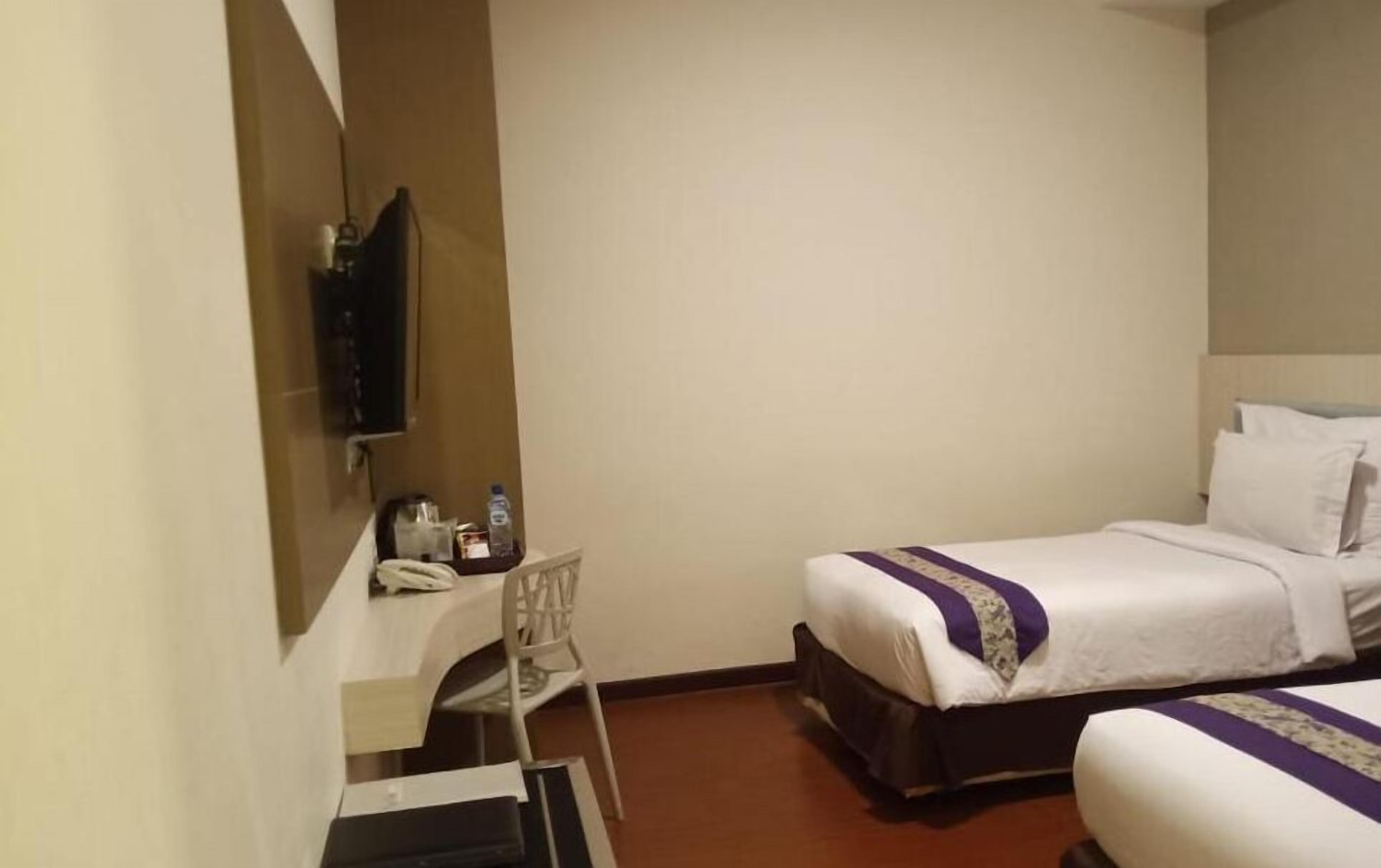 Habitación de hotel con dos camas, un escritorio con silla, un televisor y una pequeña mesa con una botella de agua y otros objetos.