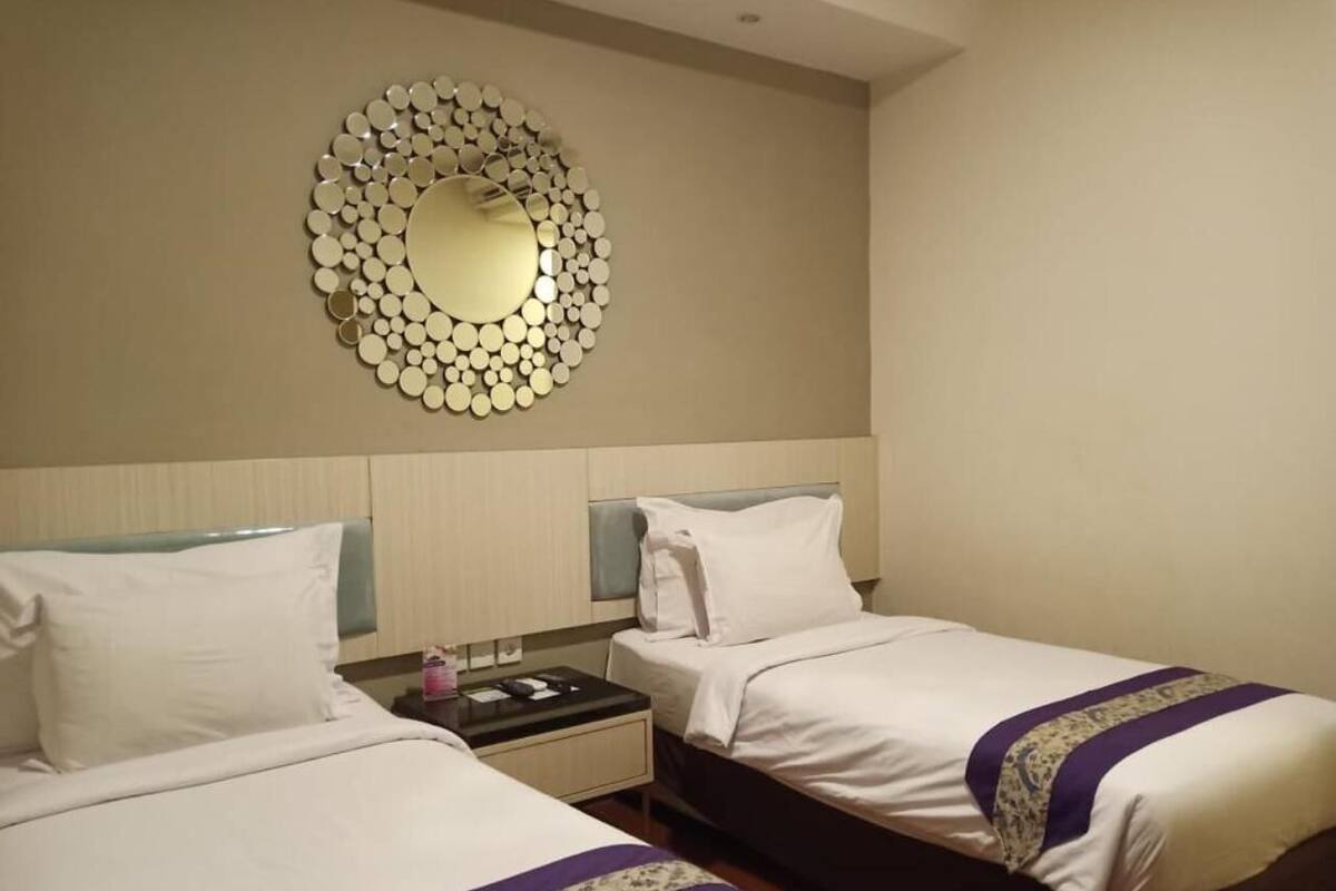 A hotel room with two beds, a large circular mirror on the wall, and a nightstand with a lamp.
