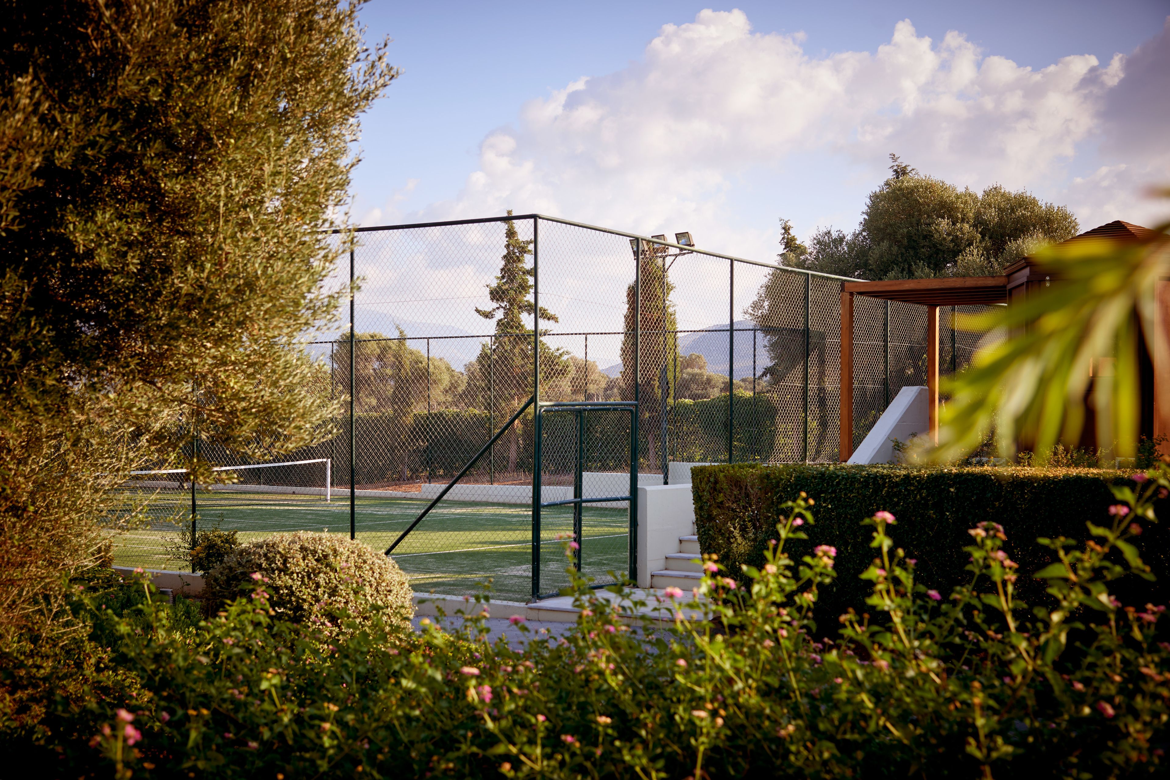 A tennis court surrounded by a fence, with a small building and a hedge in the foreground.