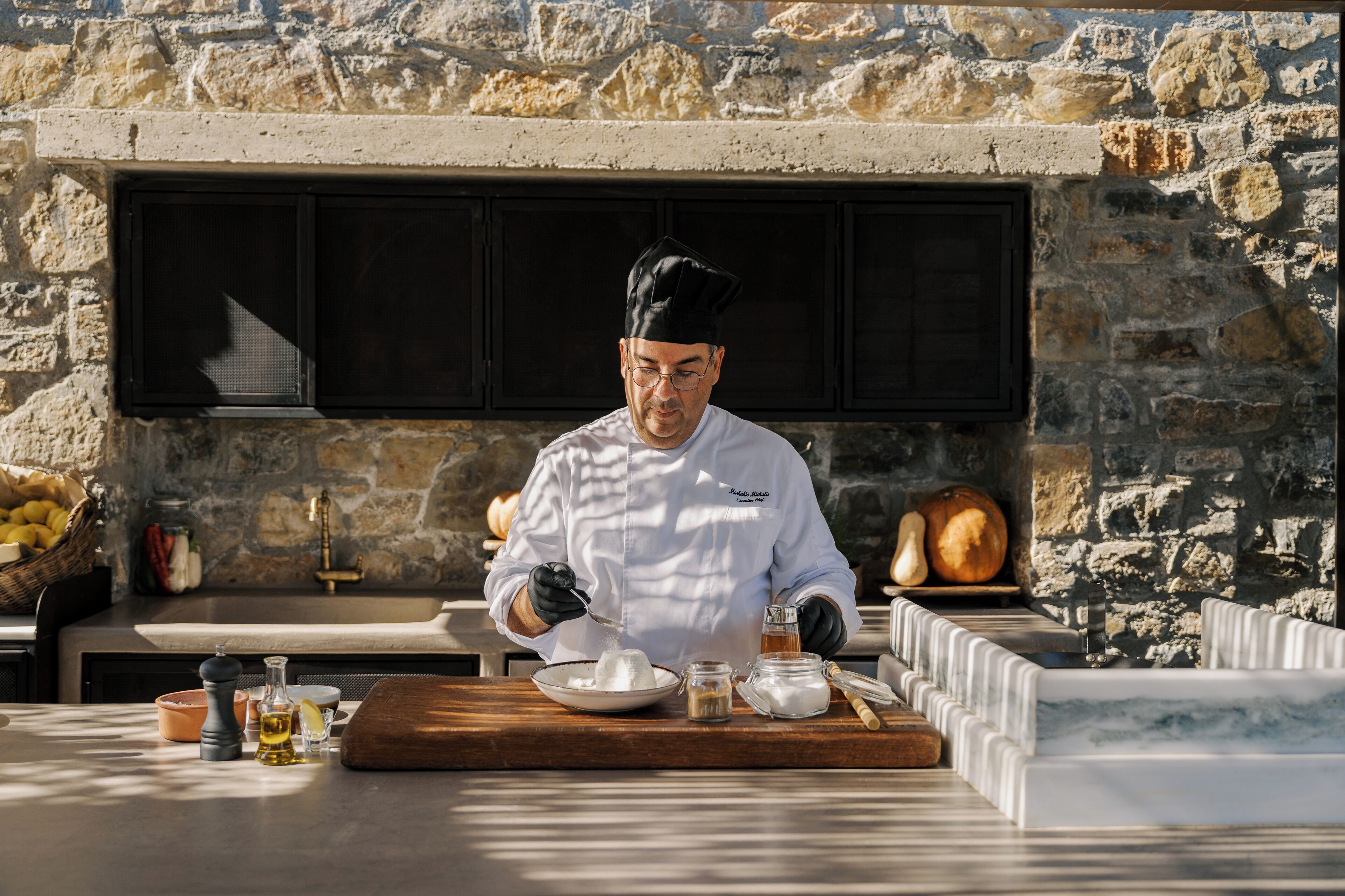 Un chef con uniforme blanco y gorro negro preparando comida en una mesa de madera, con un fondo de pared de piedra.