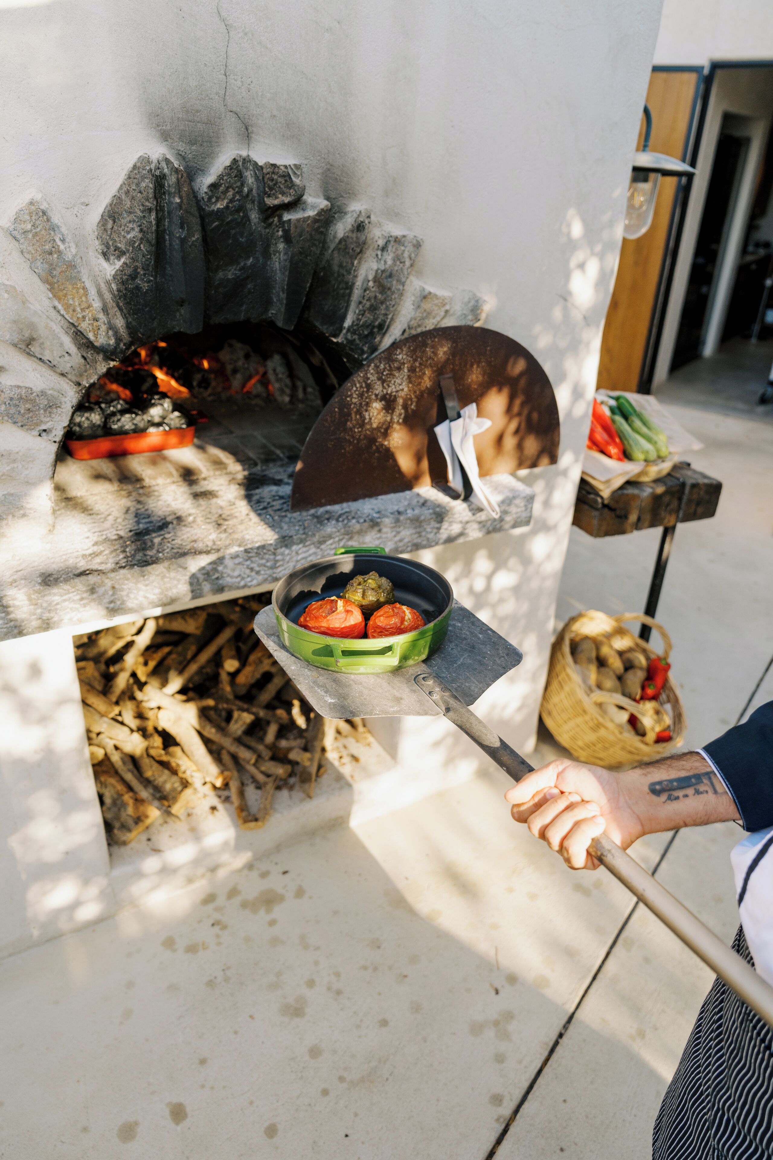 Un forno in pietra con cibo in cottura, un cesto di verdure e una persona con un tatuaggio sul braccio.