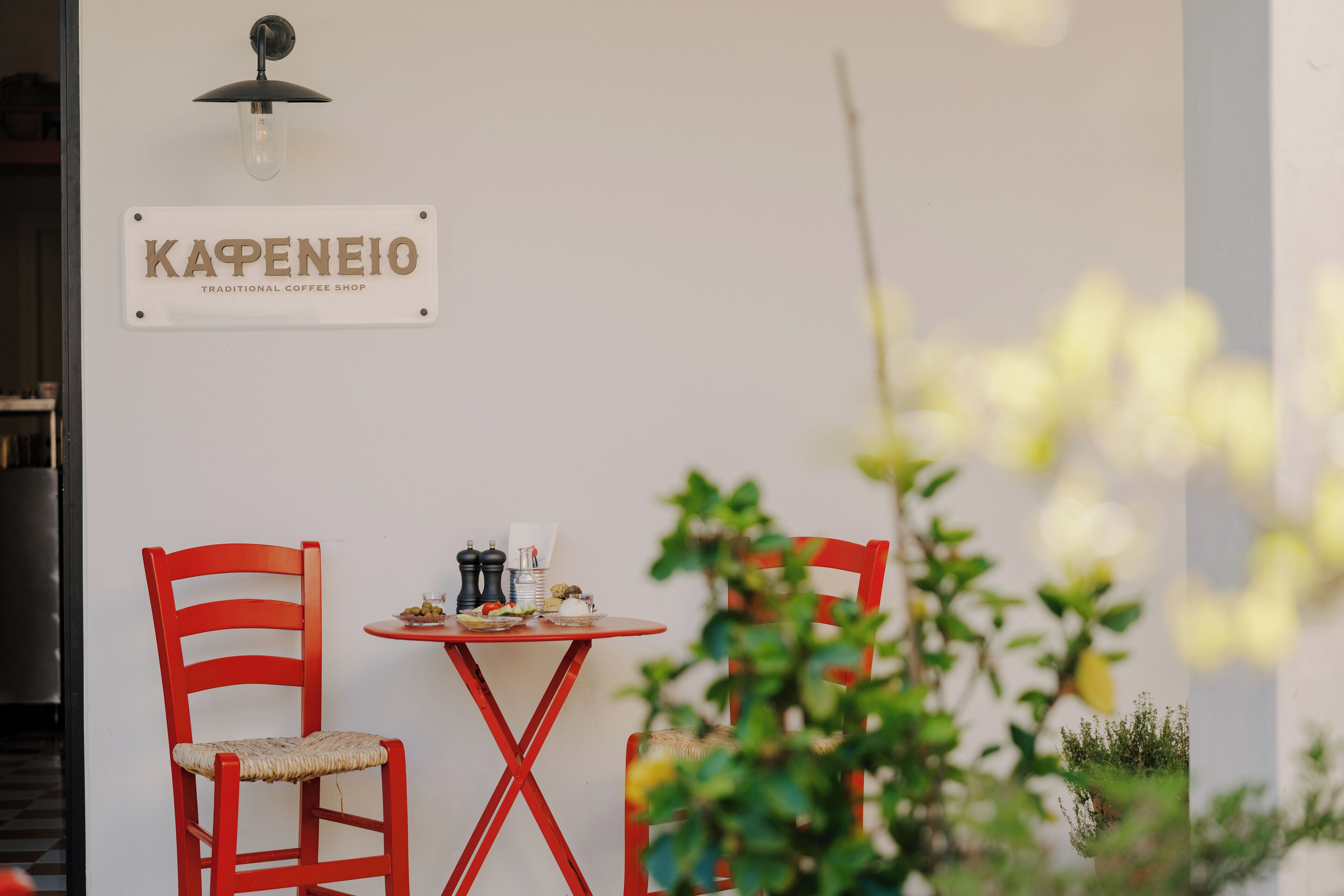 Una cafetería tradicional con un letrero que dice "Капенео". Hay dos sillas rojas y una mesa pequeña con una cafetera, azúcar y una taza.
