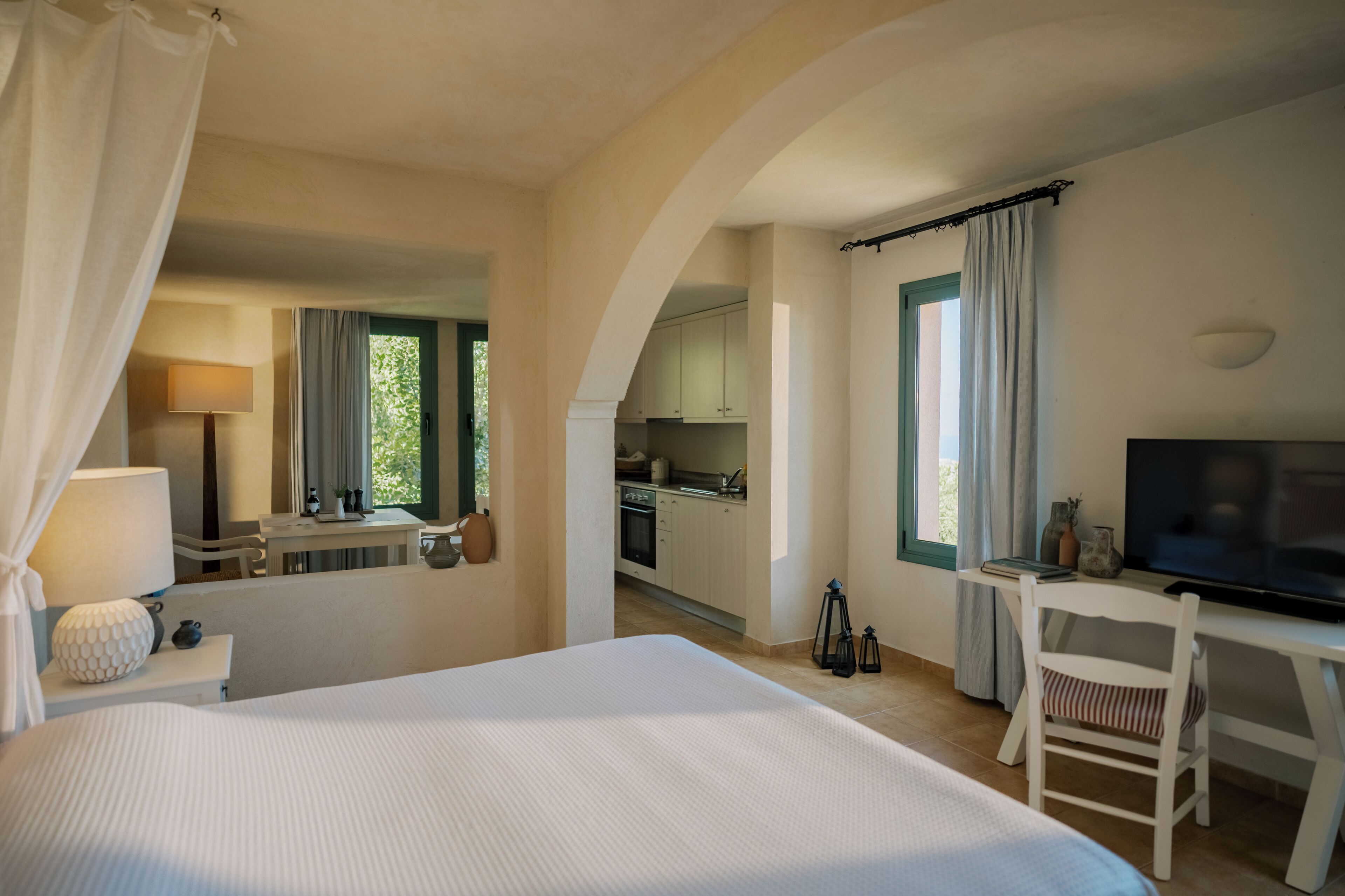 A hotel room with a large bed, a desk with a chair, a television, and a kitchenette in the background.