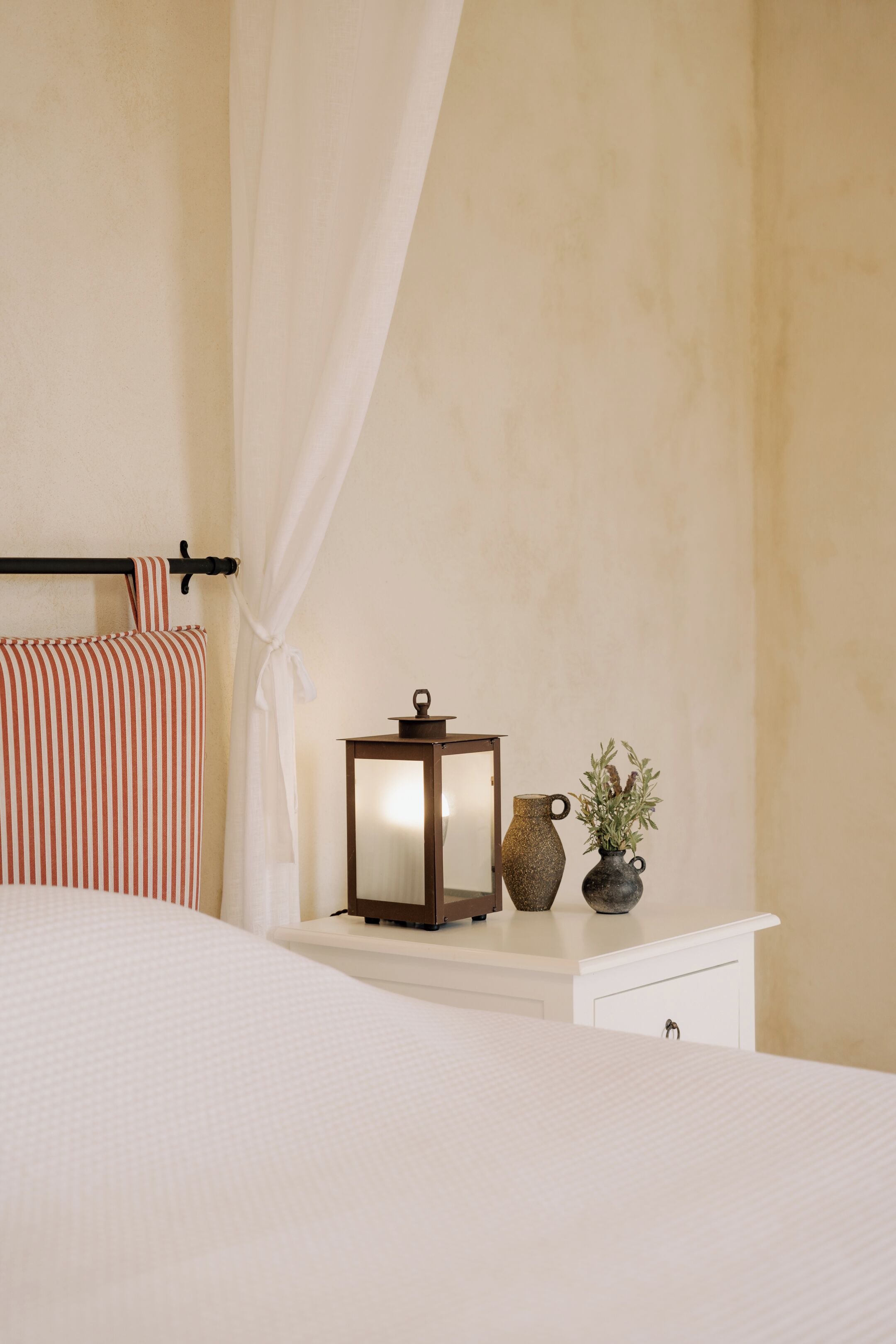 A bedroom with a bed, a bedside table with a lamp and a vase, and a curtain.