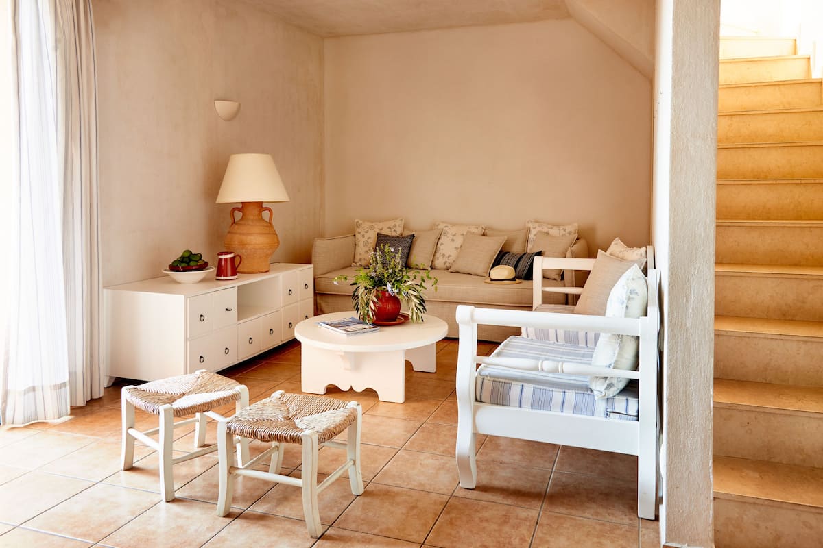 A cozy living room with a sofa, coffee table, and wicker chairs. A lamp and a vase with flowers are on the coffee table.