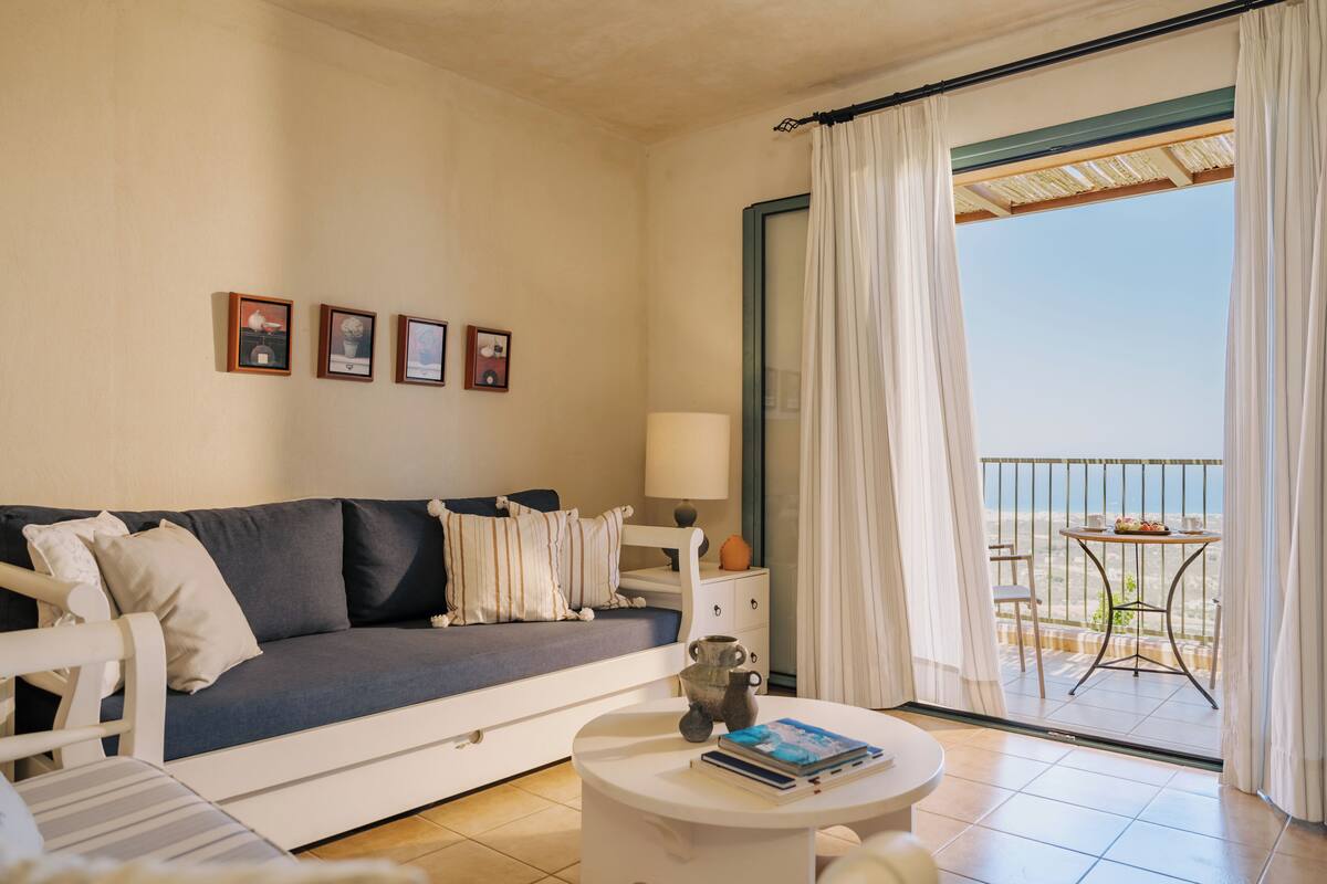 A room with a sofa, a coffee table, a lamp, and a view of the sea through a balcony.